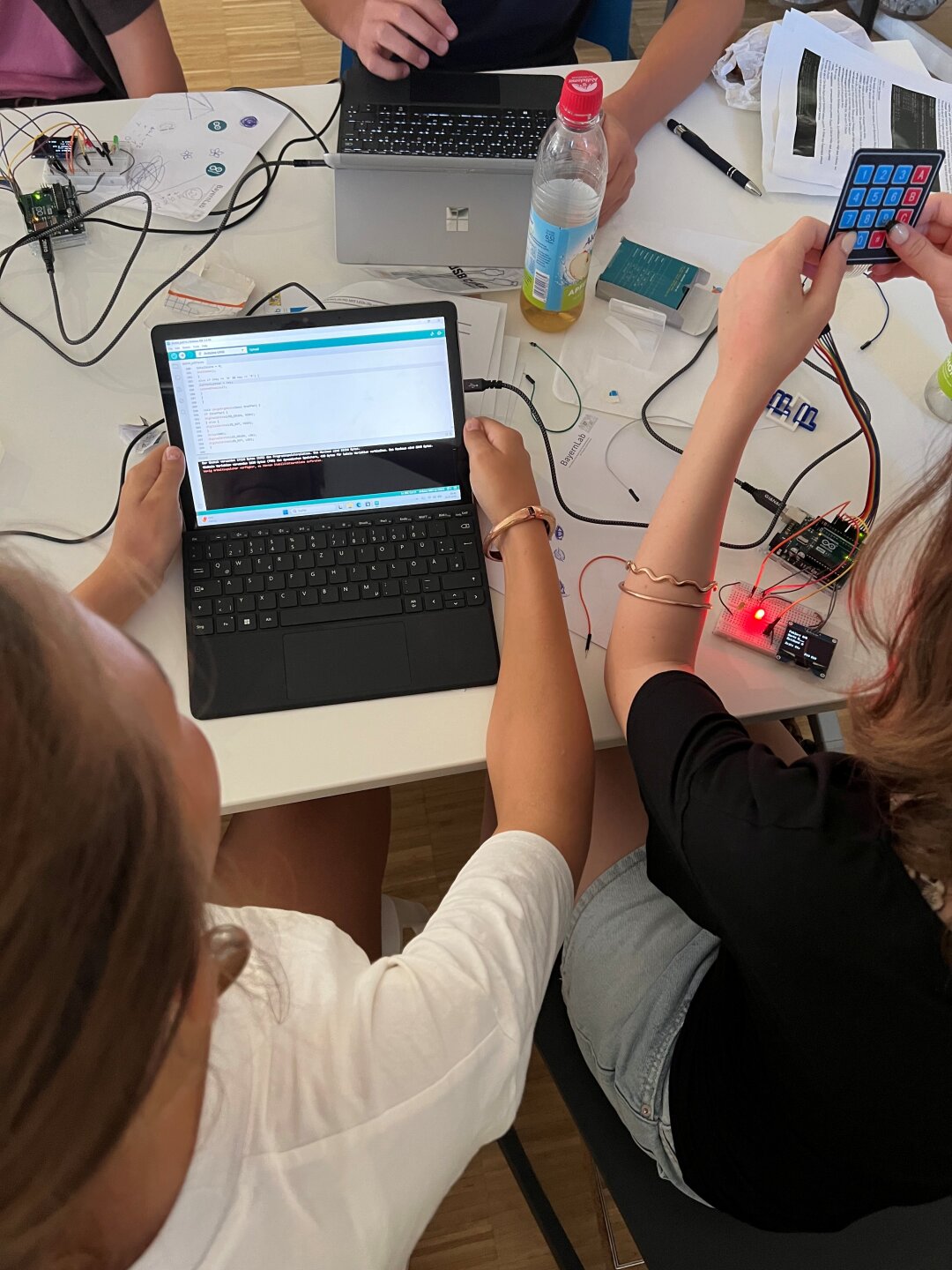 2 Schülerinnen, sie sind von hinten zu sehen, sitzen vor einem aufgeklappten Laptop, auf dem ein Code zu sehen ist. An einem Matrix-Keypad blinkt eine LED. 